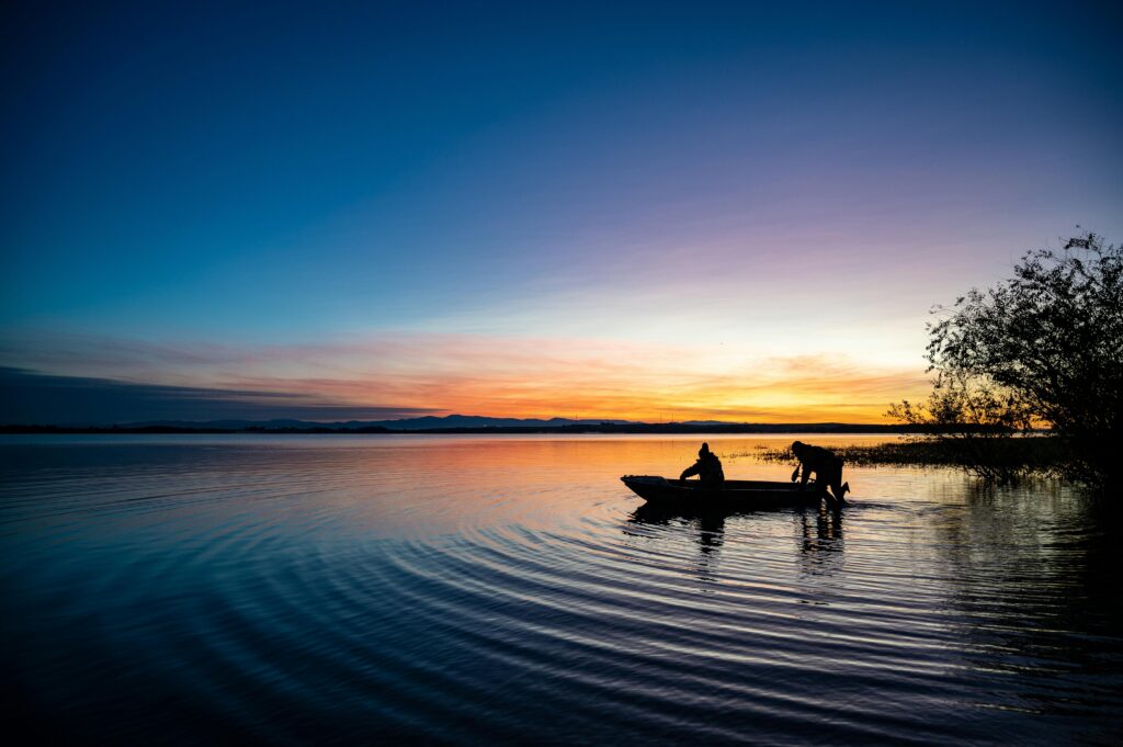 Silhouetted people in a boat at sunset on a serene lake, reflecting vibrant dusk colors.