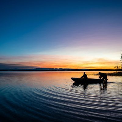 Silhouetted people in a boat at sunset on a serene lake, reflecting vibrant dusk colors.